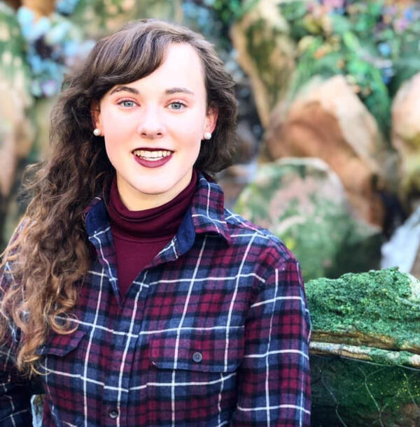 Ali, a female-presenting small human with curly, long brown hair is in front of mossy stones. She is wearing a blue and burgundy plaid shirt with a burgundy turtleneck underneath. She is smiling and has burgundy lipstick on as well as pearl earrings.